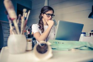 Woman sits in front of a computer on a messy desk with her chin in her hand and a concerned look on her face