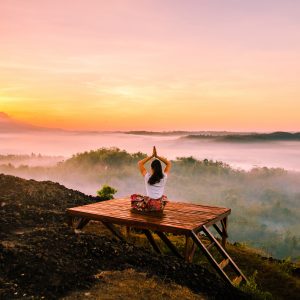 Woman with hands overhead in prayer pose sitting on a platform overlooking a scenic valley at sunset