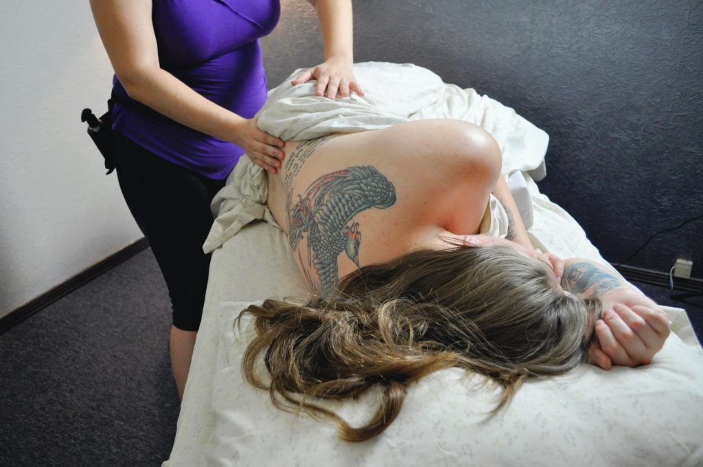 A woman performs a prenatal massage on a pregnant woman who is lying on her side propped up by pillows.